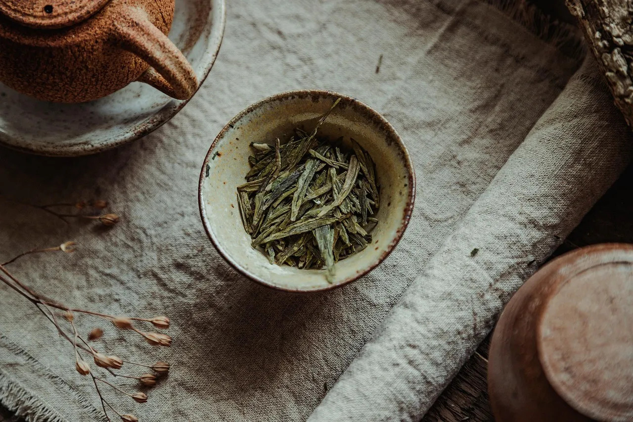 Flache grüne Longjing-Teeblätter in einer kleinen Schale neben einer Teekanne