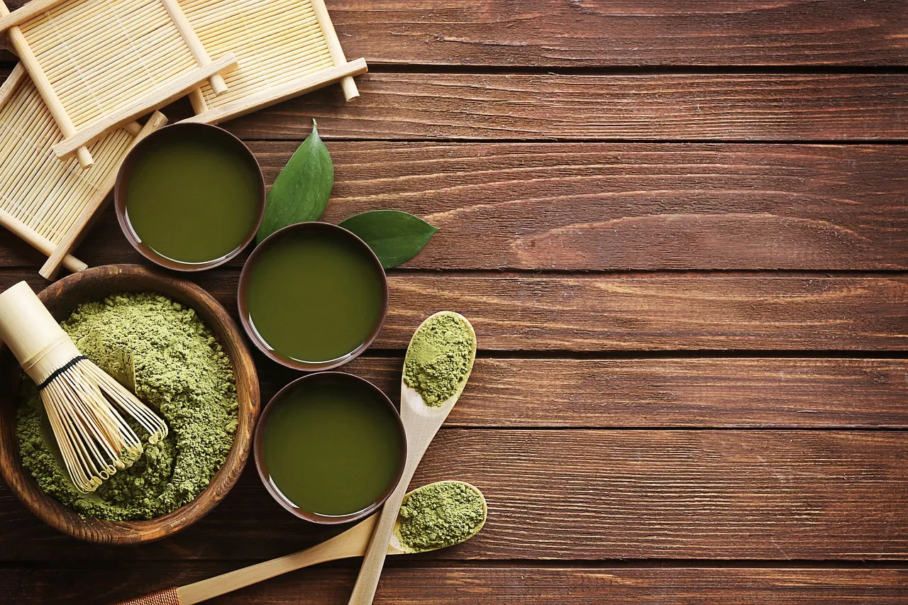 Matcha-Pulver, ein Bambusbesen und Schalen mit aufgeschlagenem Matcha auf einem Holztisch